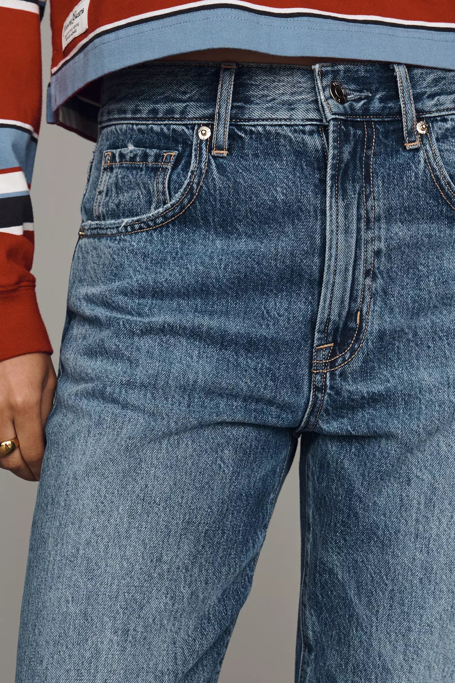 A model stands against a solid gray studio background wearing a cropped, long-sleeve striped knit polo shirt in rust red, navy, white, and light blue, paired with wide-leg blue jeans