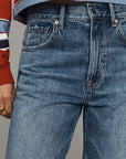 A model stands against a solid gray studio background wearing a cropped, long-sleeve striped knit polo shirt in rust red, navy, white, and light blue, paired with wide-leg blue jeans