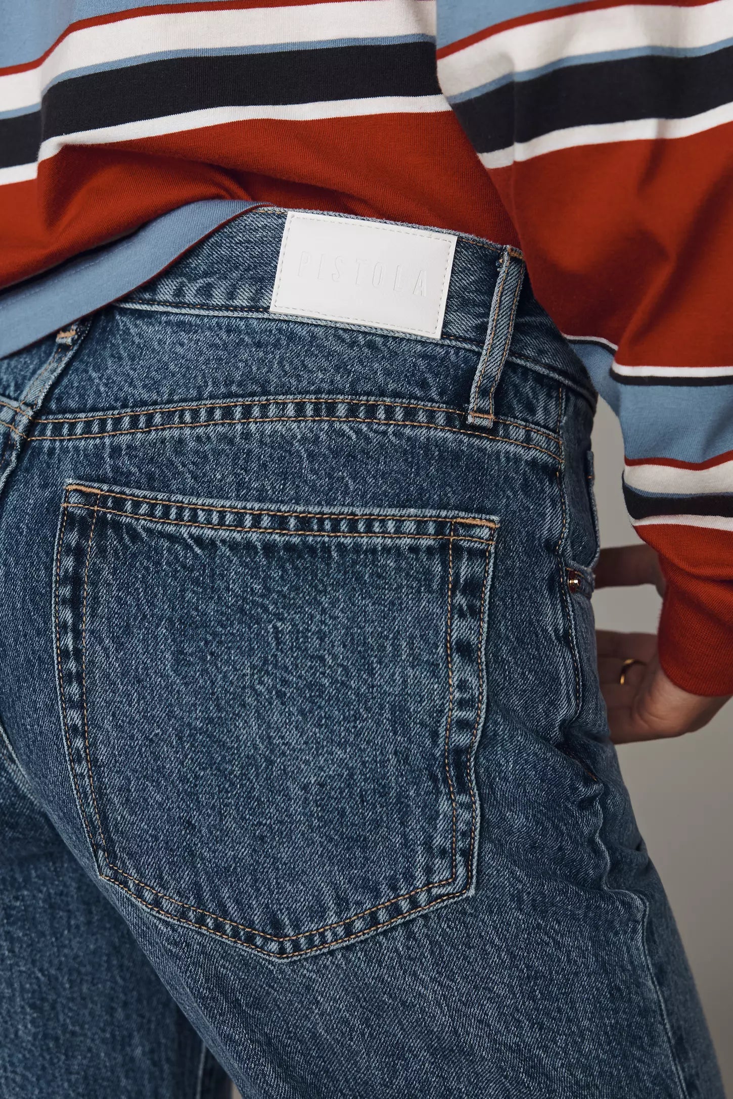 Close-up of the back waistband and pocket area of blue denim jeans, worn with a striped long-sleeve shirt. A blank white rectangular patch is visible at the back waistband.