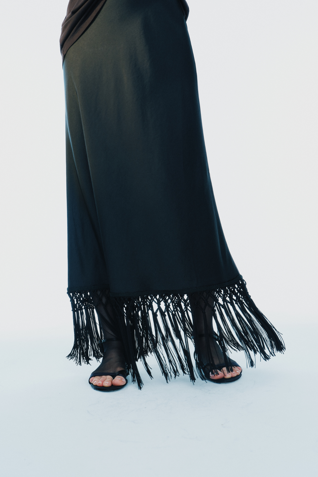 A black maxi skirt with a fringed hem, shown on a model wearing black strappy flat sandals against a plain white studio background.