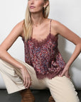 A woman crouching in a studio wears a burgundy floral lace camisole top with thin straps, paired with cream pants and brown ankle boots against a light gray background.