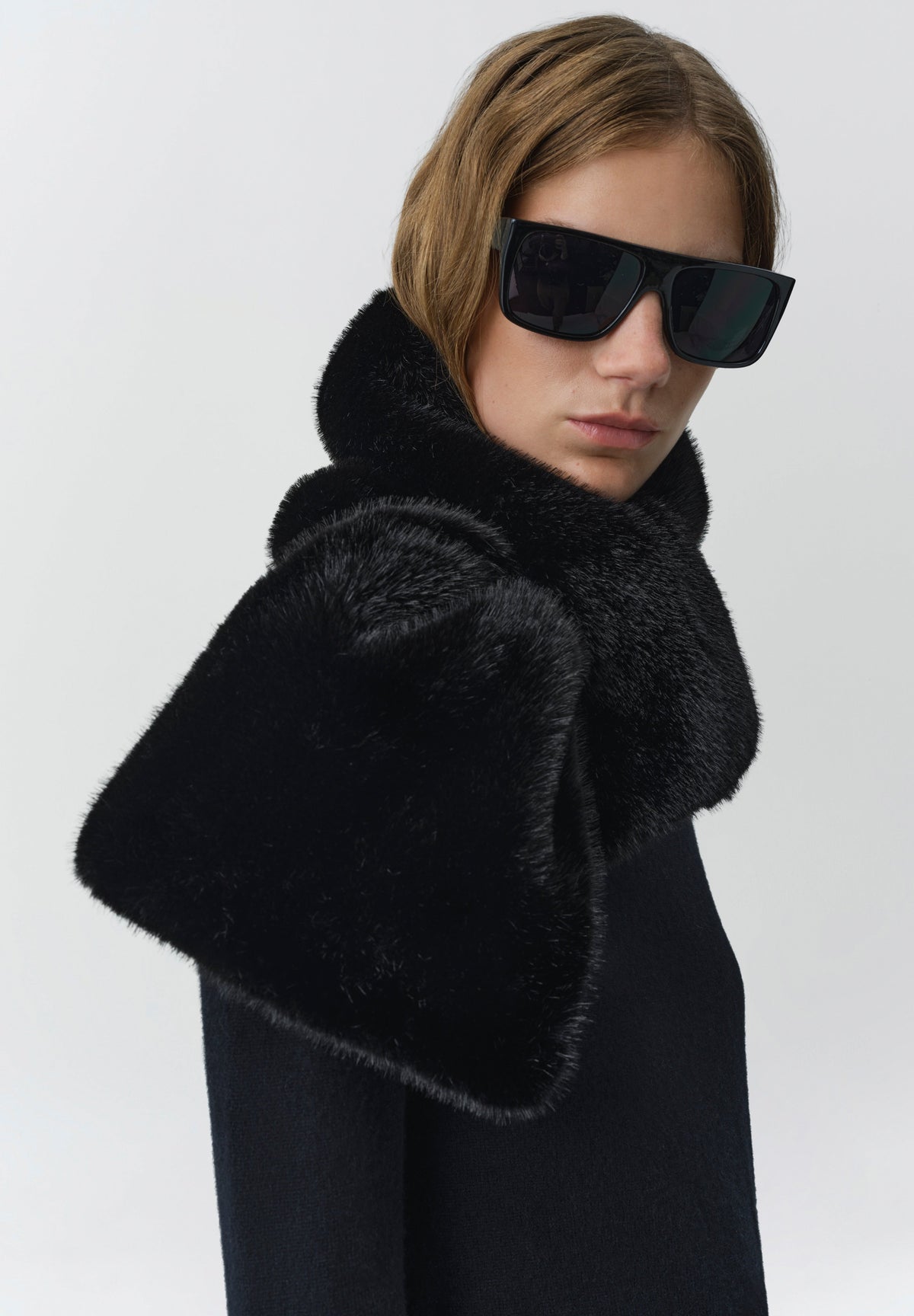 A model wearing a luxurious black fur collar with wide flared cuffs and black sunglasses, set against a plain light gray studio background.