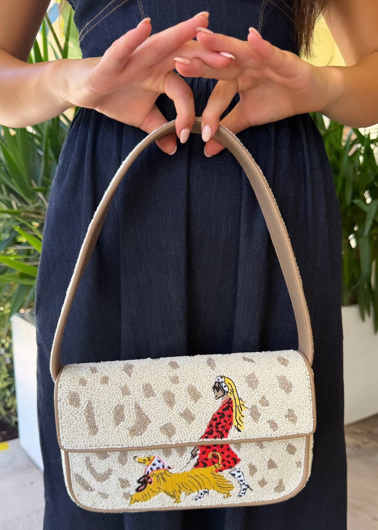 A person in a navy dress holds a New Staud Tommy beaded handbag with a leopard print pattern and a colorful embroidered figure on the front flap.