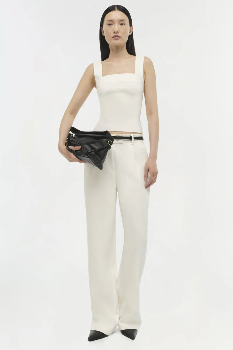 Woman wearing a white sleeveless top and pants with a black clutch against a white background