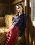 A woman leans against a wooden wall in a barn, wearing a navy and red striped long-sleeve polo shirt with a dark collar and a long, flowing dusty rose skirt.