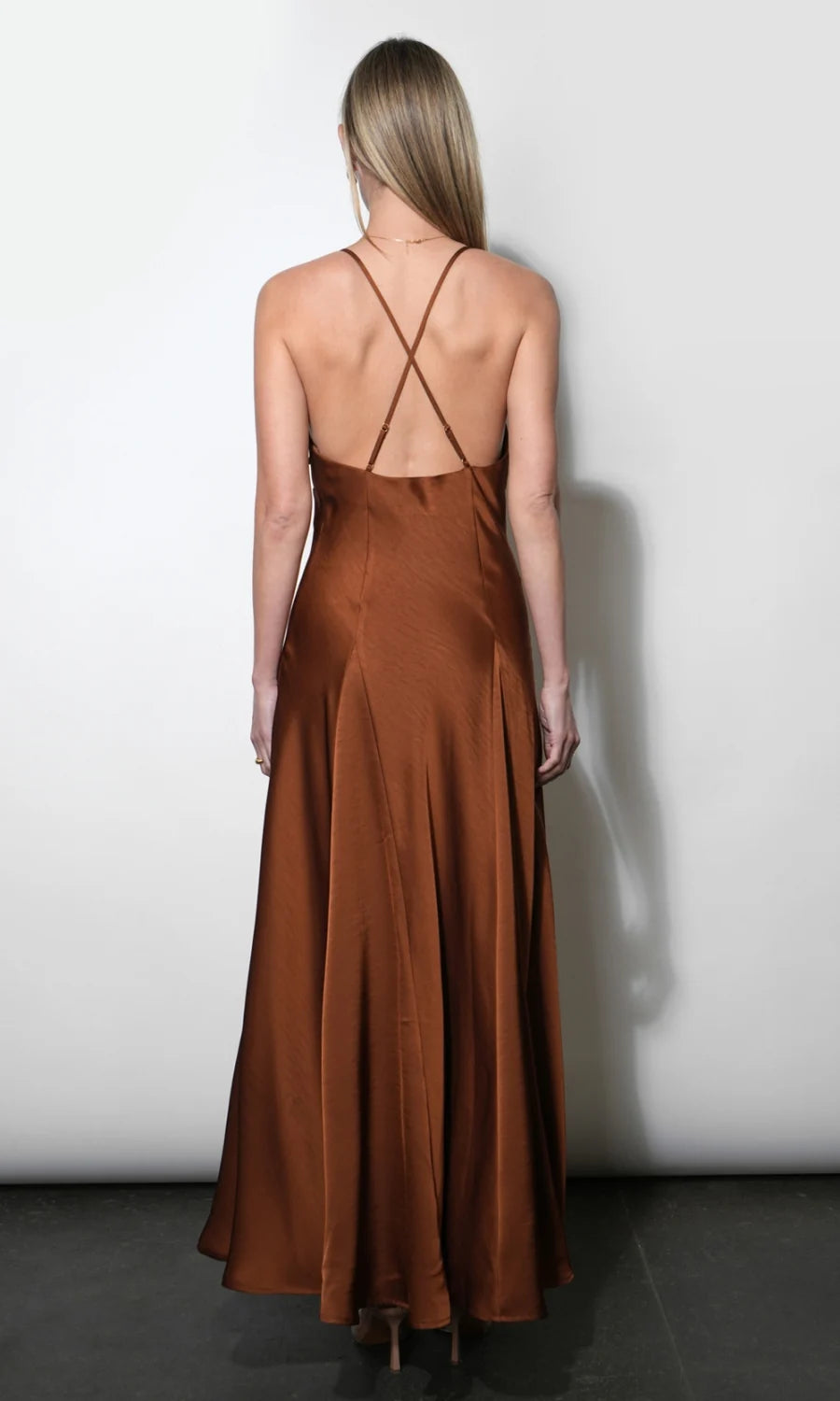 A woman viewed from the back wearing a long satin-like slip dress in a warm rust brown color, with thin crisscross back straps, standing against a plain light gray wall.