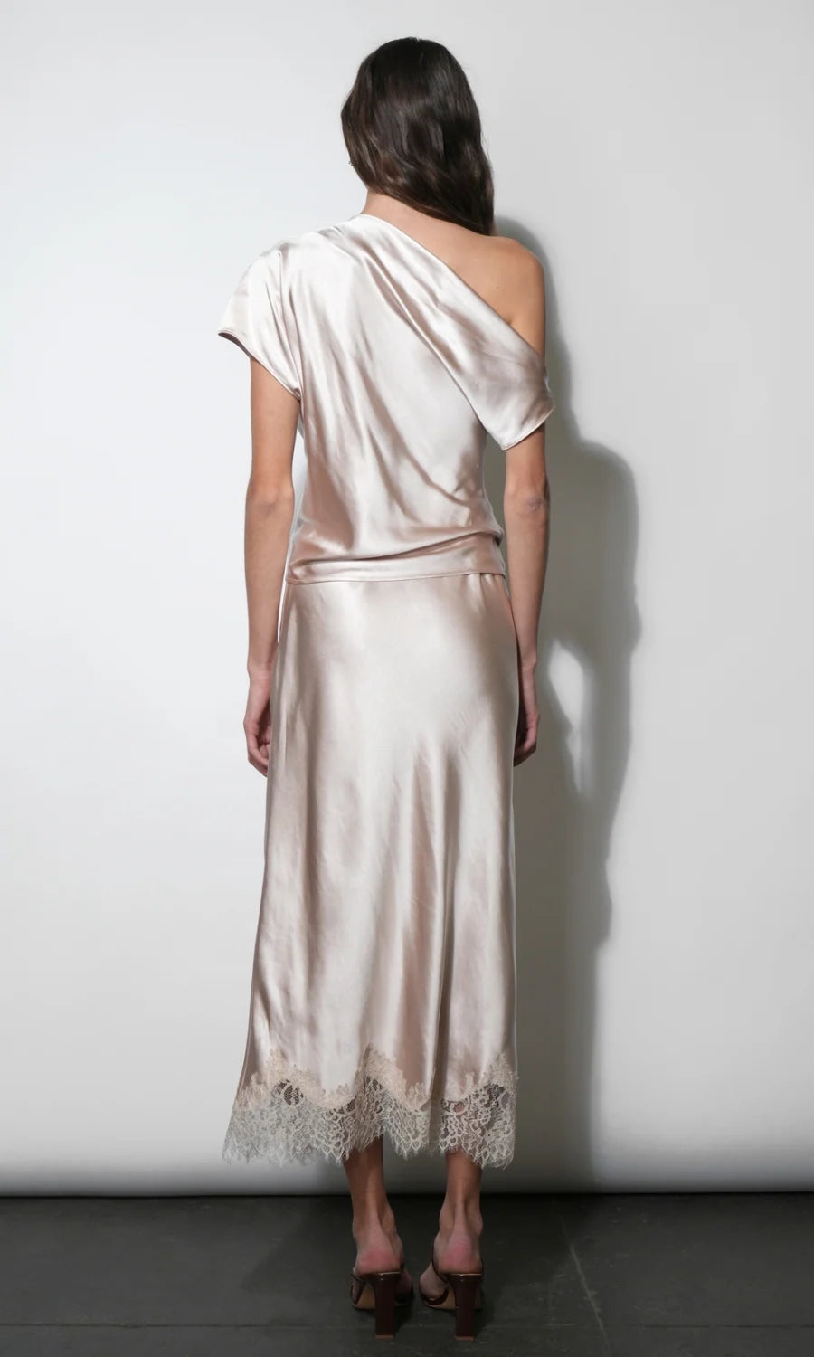 A woman viewed from behind wearing a satin one-shoulder midi dress in a pale blush pink color, standing against a plain light gray wall.