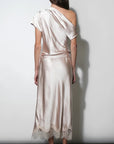 A woman viewed from behind wearing a satin one-shoulder midi dress in a pale blush pink color, standing against a plain light gray wall.