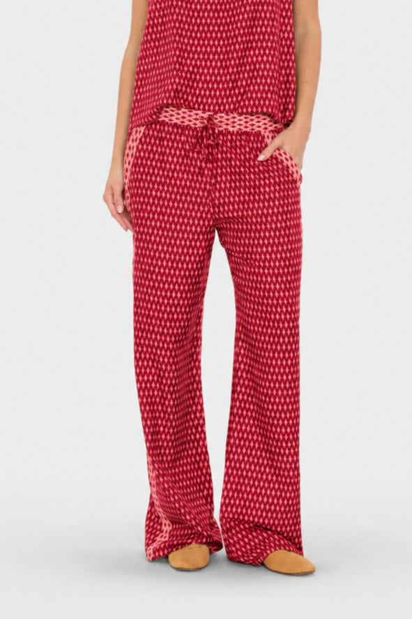 A person wearing red and white patterned lounge pants with a matching drawstring belt and a matching red top, standing against a light gray studio background.