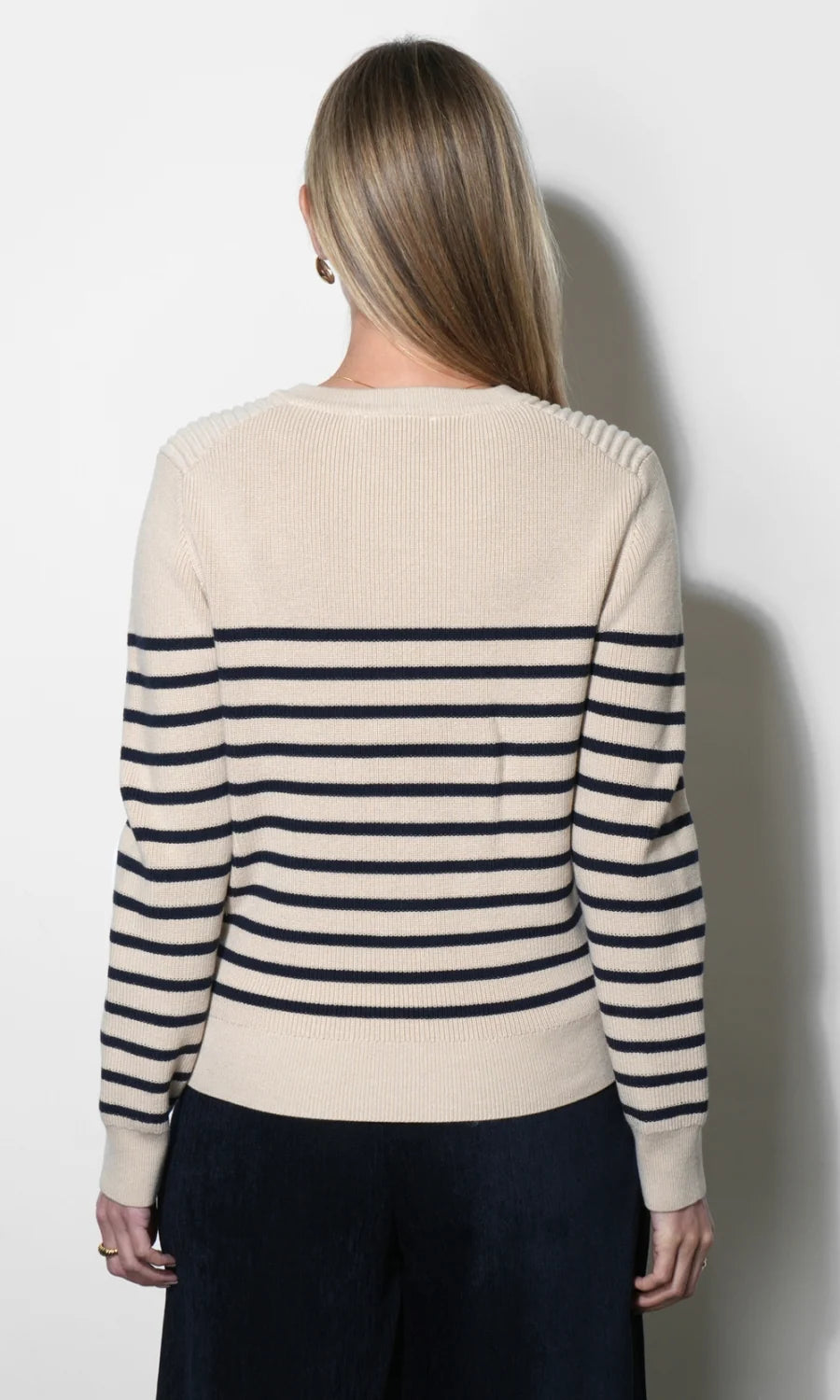 A woman viewed from the back wearing a cream knit sweater with navy horizontal stripes across the shoulders and sleeves, paired with dark navy pants, against a plain light gray wall.