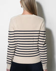 A woman viewed from the back wearing a cream knit sweater with navy horizontal stripes across the shoulders and sleeves, paired with dark navy pants, against a plain light gray wall.
