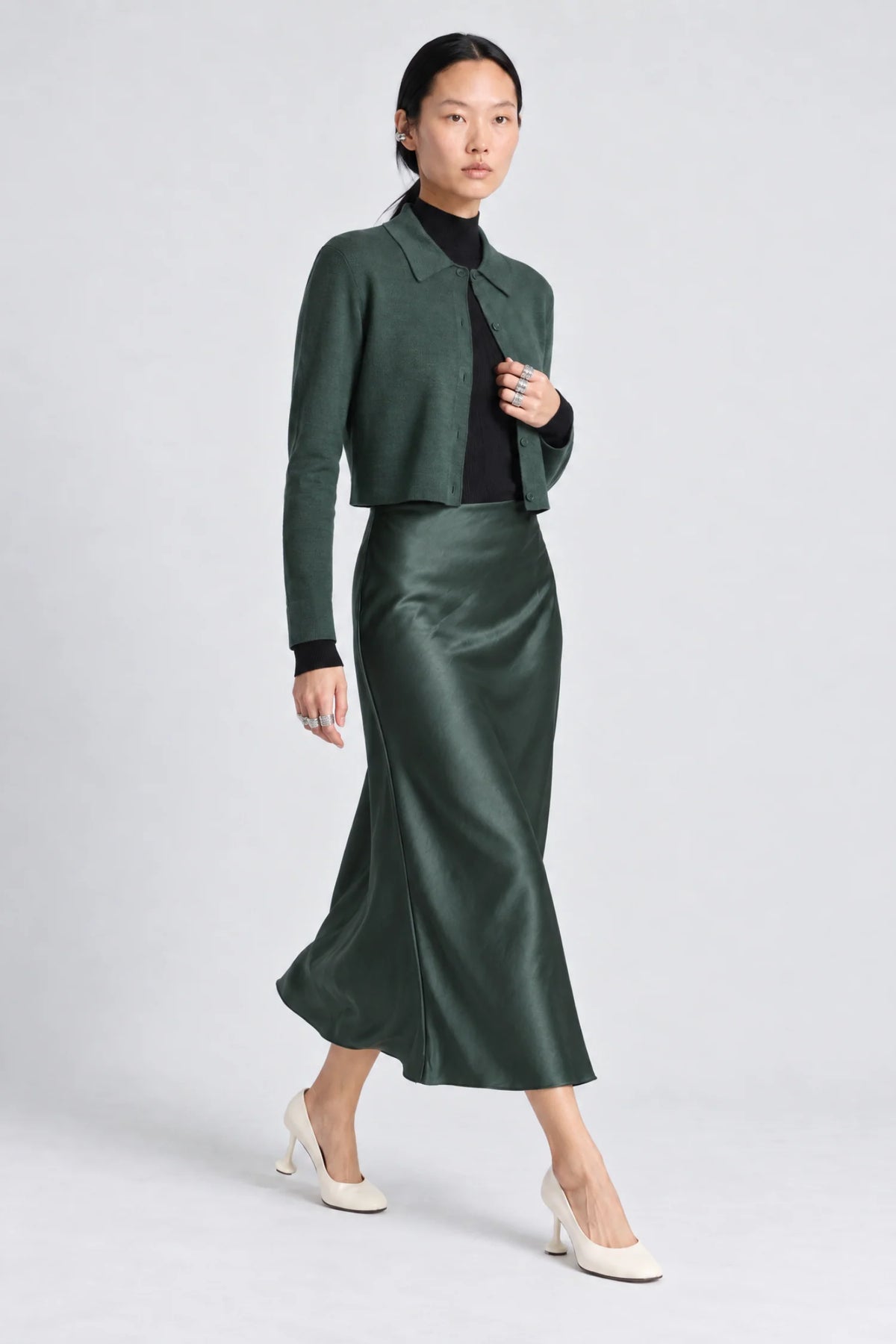 A model wearing a dark green cropped sweater and matching satin midi skirt with a black turtleneck and white heels, standing against a light gray studio background.
