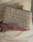 A cream-colored fringe clutch bag with a gold-tone frame and two round clasp buttons, held in a hand wearing a matching cream dress and a brown bangle.