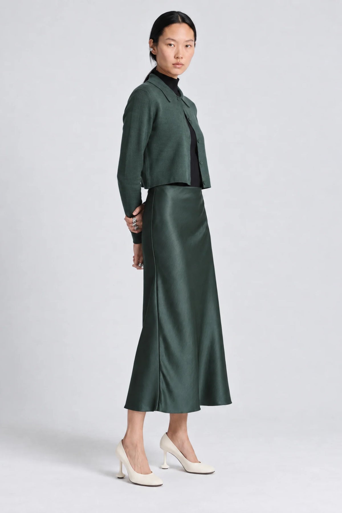 A model is shown from the side wearing a dark green set consisting of a cropped sweater and a midi skirt, paired with white high heels, against a light gray studio background