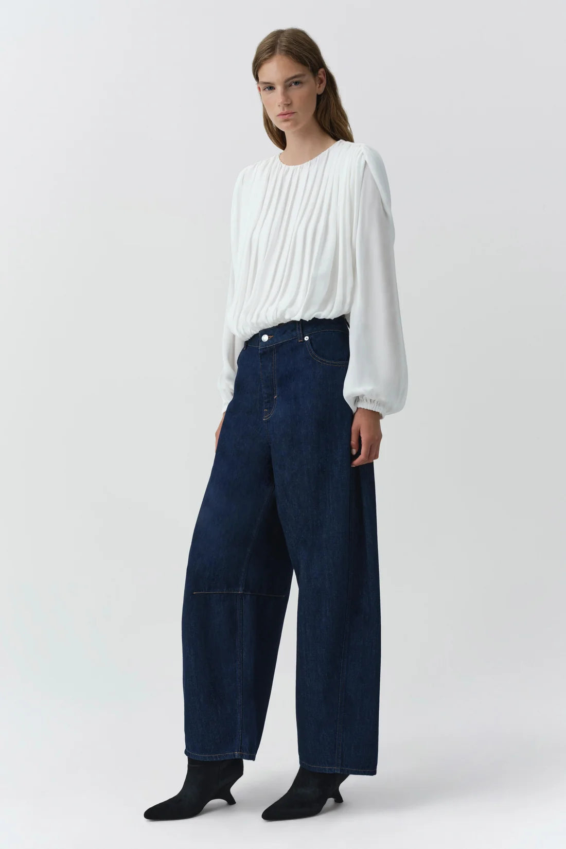 A model wearing a white long-sleeve pleated blouse and dark blue wide-leg denim jeans, paired with black ankle boots, against a light gray studio background.