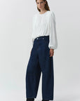 A model wearing a white long-sleeve pleated blouse and dark blue wide-leg denim jeans, paired with black ankle boots, against a light gray studio background.