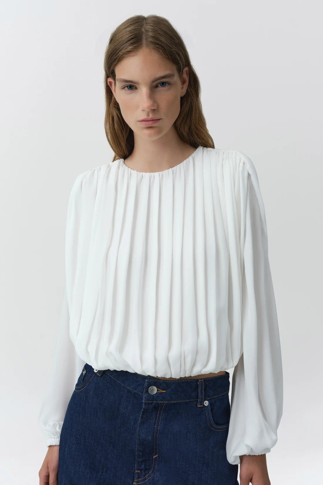 A woman wearing a white long-sleeve blouse with vertical pleats and dark blue jeans, photographed against a light gray studio background.