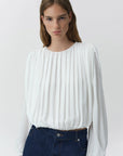 A woman wearing a white long-sleeve blouse with vertical pleats and dark blue jeans, photographed against a light gray studio background.