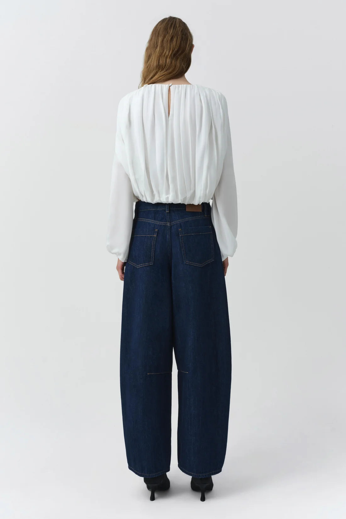 A woman viewed from the back wearing a white long-sleeve pleated blouse and dark blue wide-leg denim pants, standing against a light gray studio background.