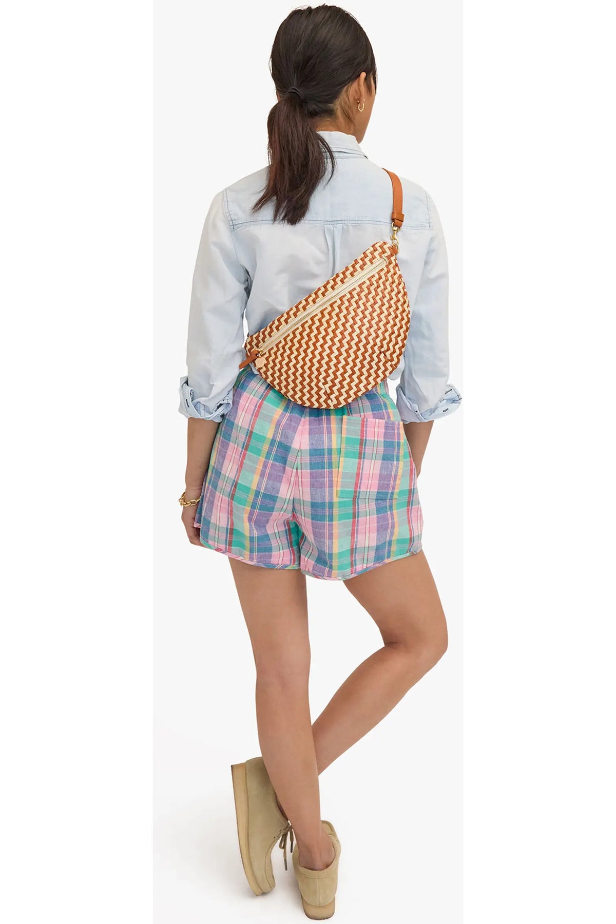 A woman shown from behind wearing a light blue denim shirt, pastel plaid shorts, and beige wedge sandals, with a woven orange and white crescent-shaped crossbody bag worn across her back.