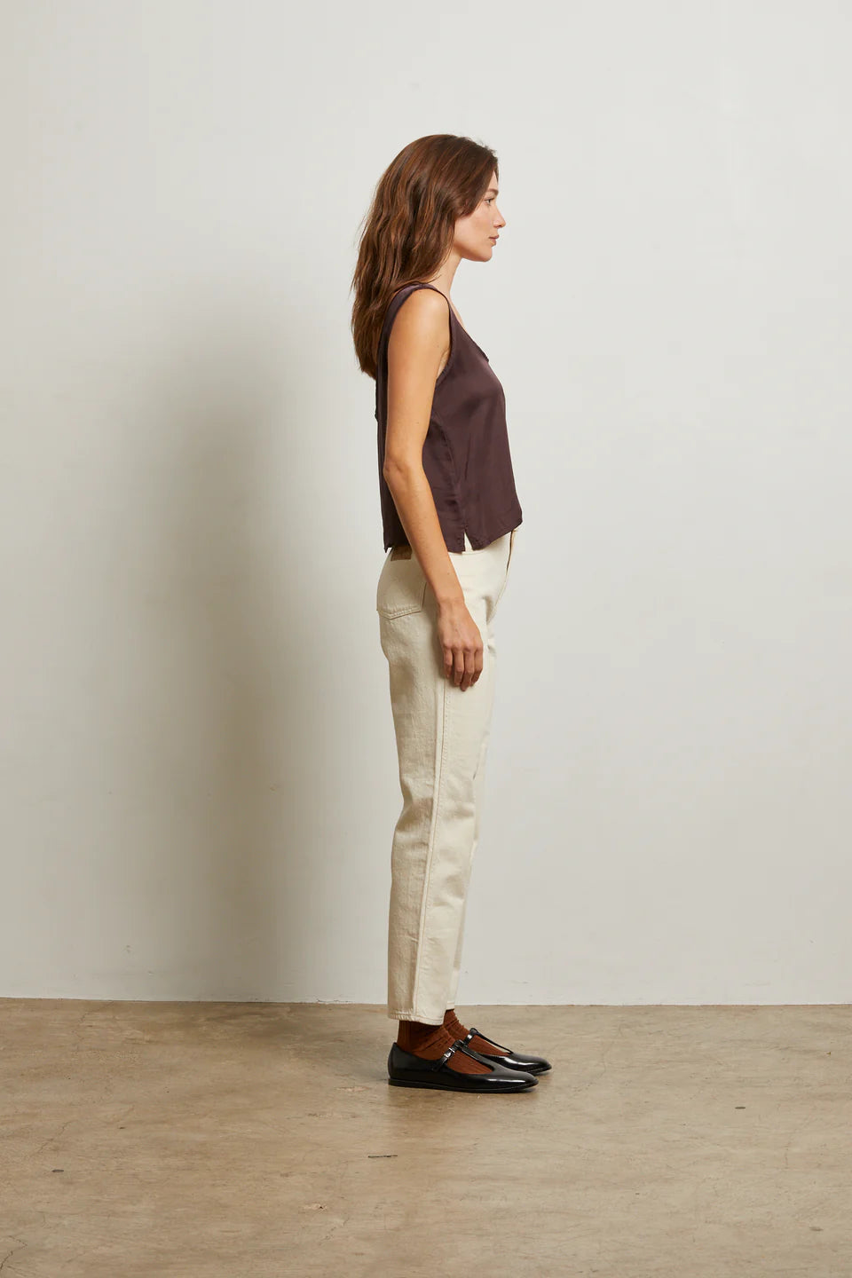 Woman standing against a plain wall wearing a sleeveless top, beige pants, and brown shoes side view
