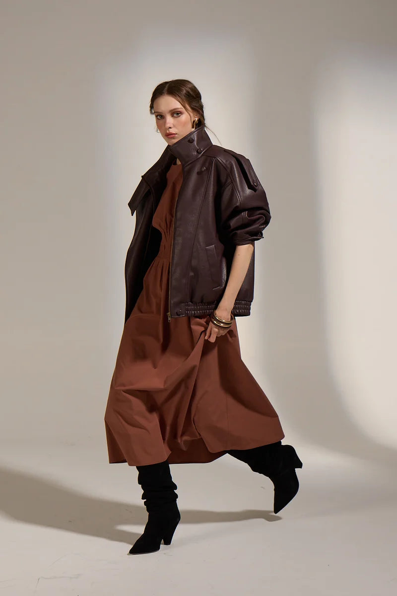 Woman wearing a brown leather jacket and dress with black boots against a neutral background