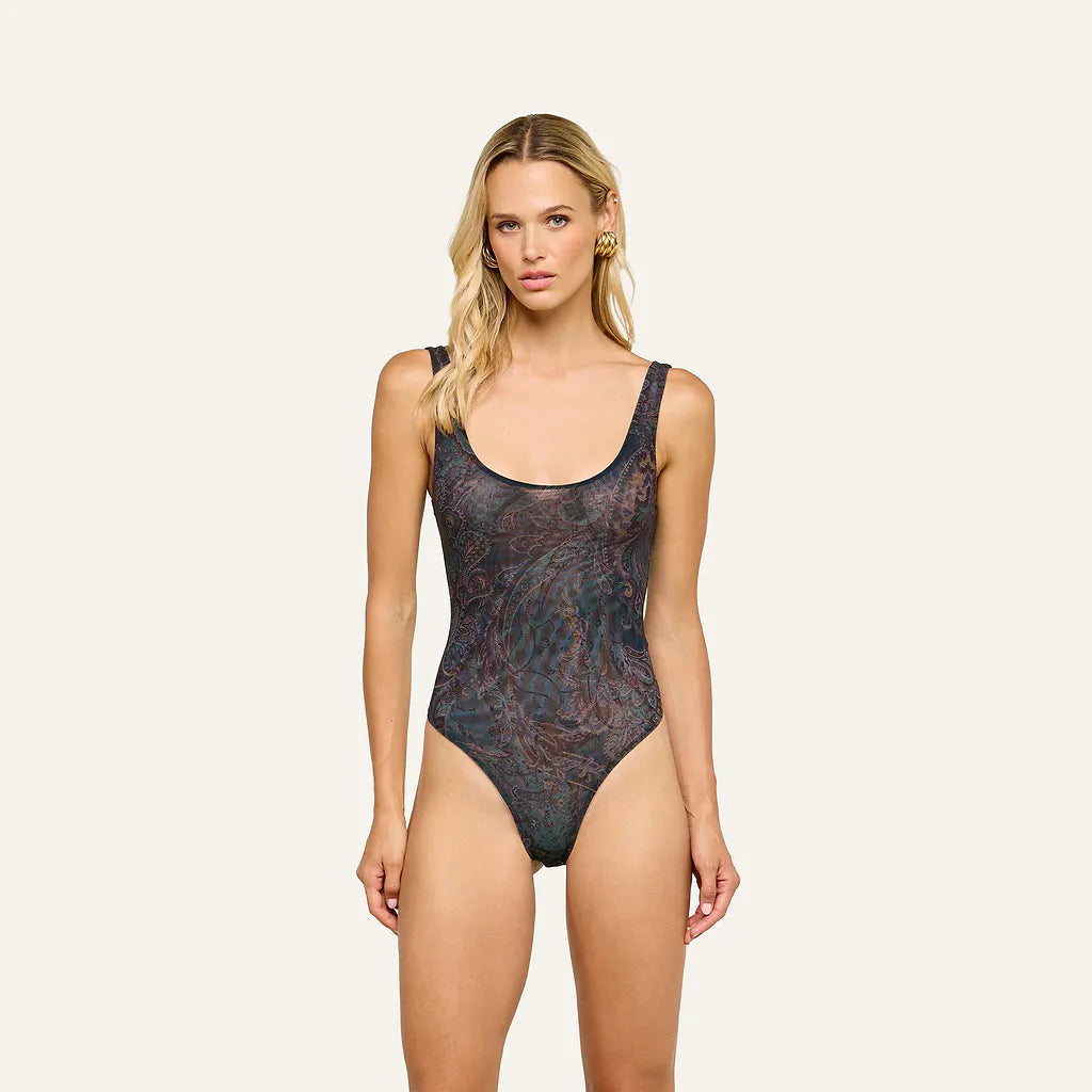 A blonde model wearing a bodysuit with a dark, abstract marbled pattern in black, blue, and brown tones, posed against a solid off-white background.