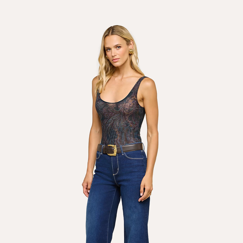 A blonde model wearing a sheer black lace bodysuit with a dark floral pattern, tucked into dark blue jeans with a brown belt, against a solid off-white background.