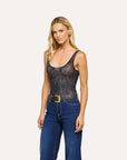A blonde model wearing a sheer black lace bodysuit with a dark floral pattern, tucked into dark blue jeans with a brown belt, against a solid off-white background.