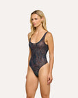 A blonde model wearing a one-piece swimsuit with a dark navy and black abstract pattern, standing against a solid off-white background.