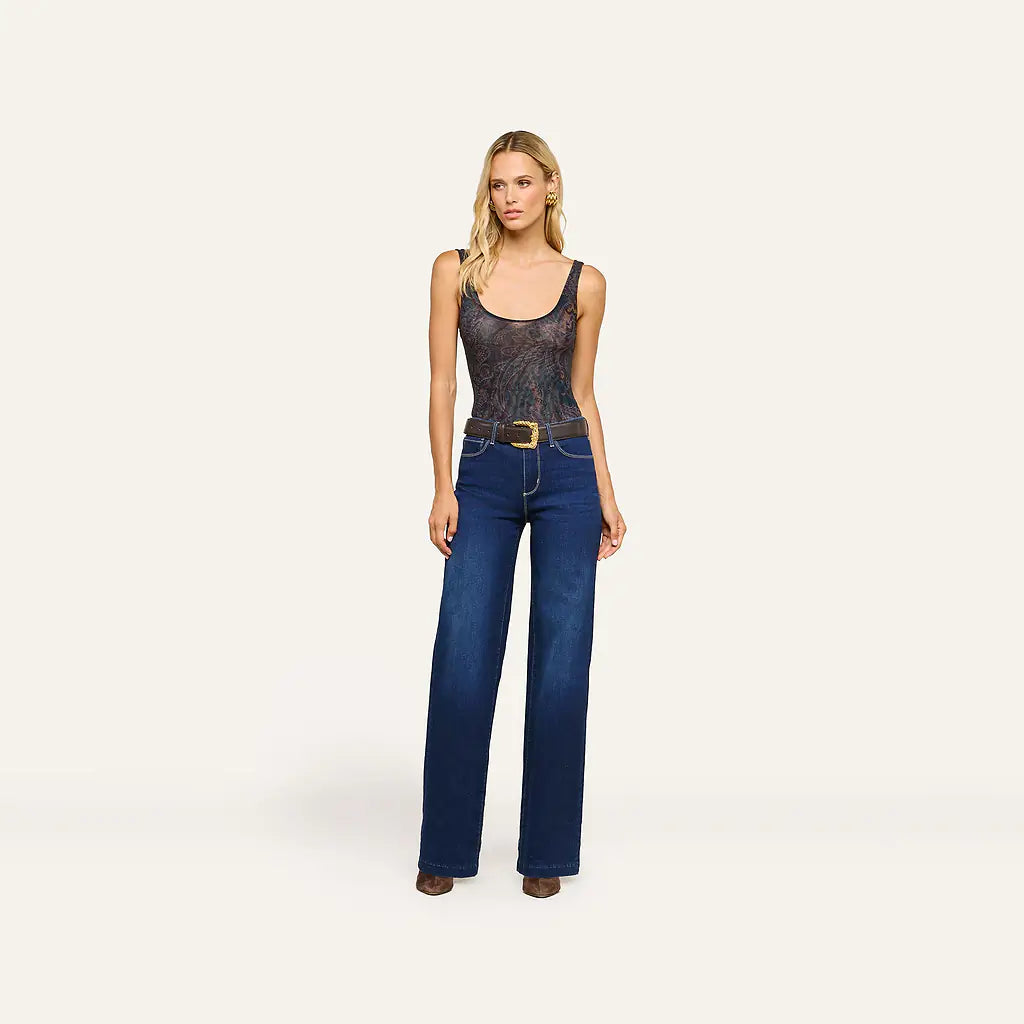 A woman stands against a plain light beige studio background wearing a sleeveless dark patterned top with a gold belt and dark blue wide-leg jeans.