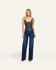 A woman stands against a plain light beige studio background wearing a sleeveless dark patterned top with a gold belt and dark blue wide-leg jeans.