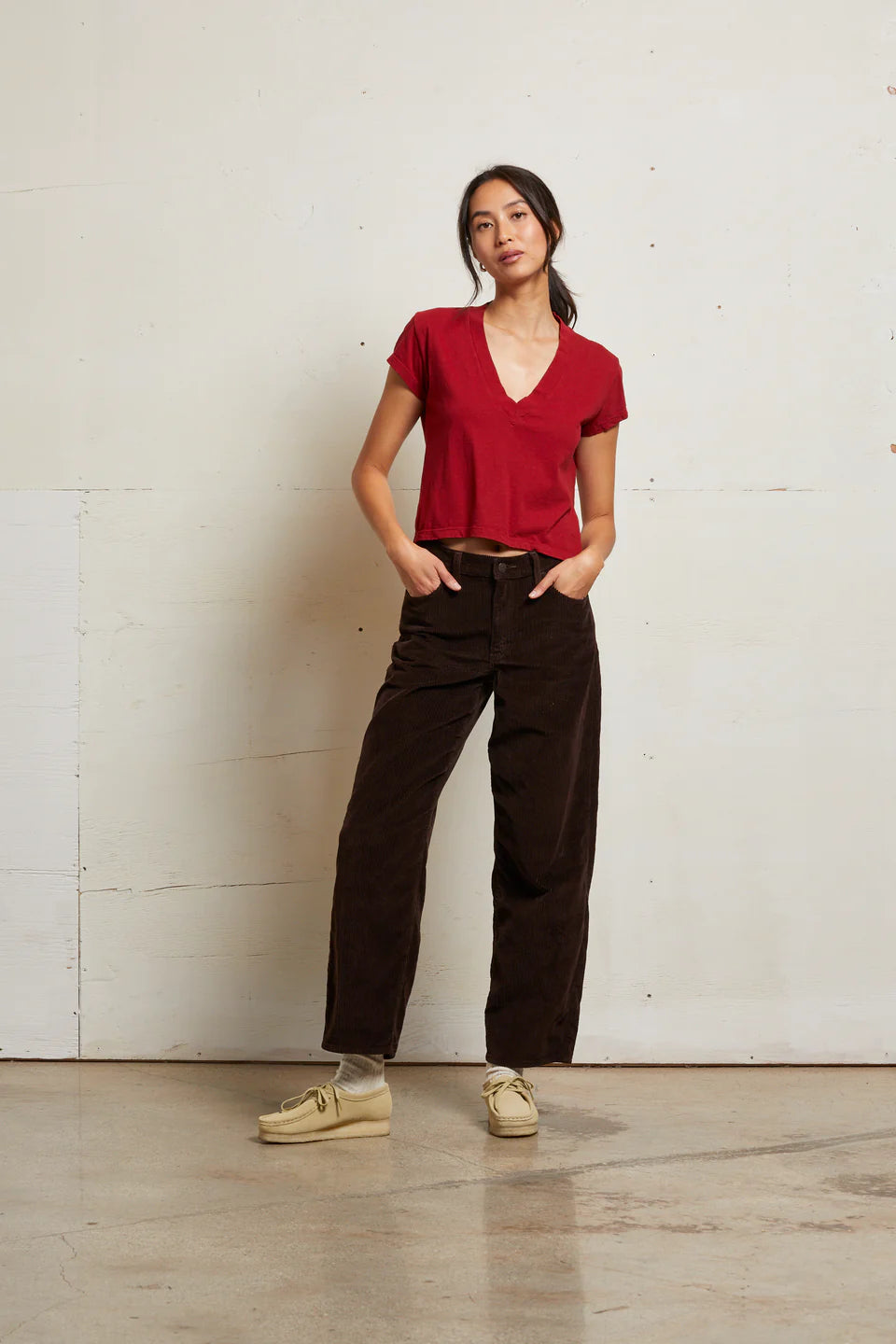 Person wearing a red top and brown pants standing against a white wall. front view