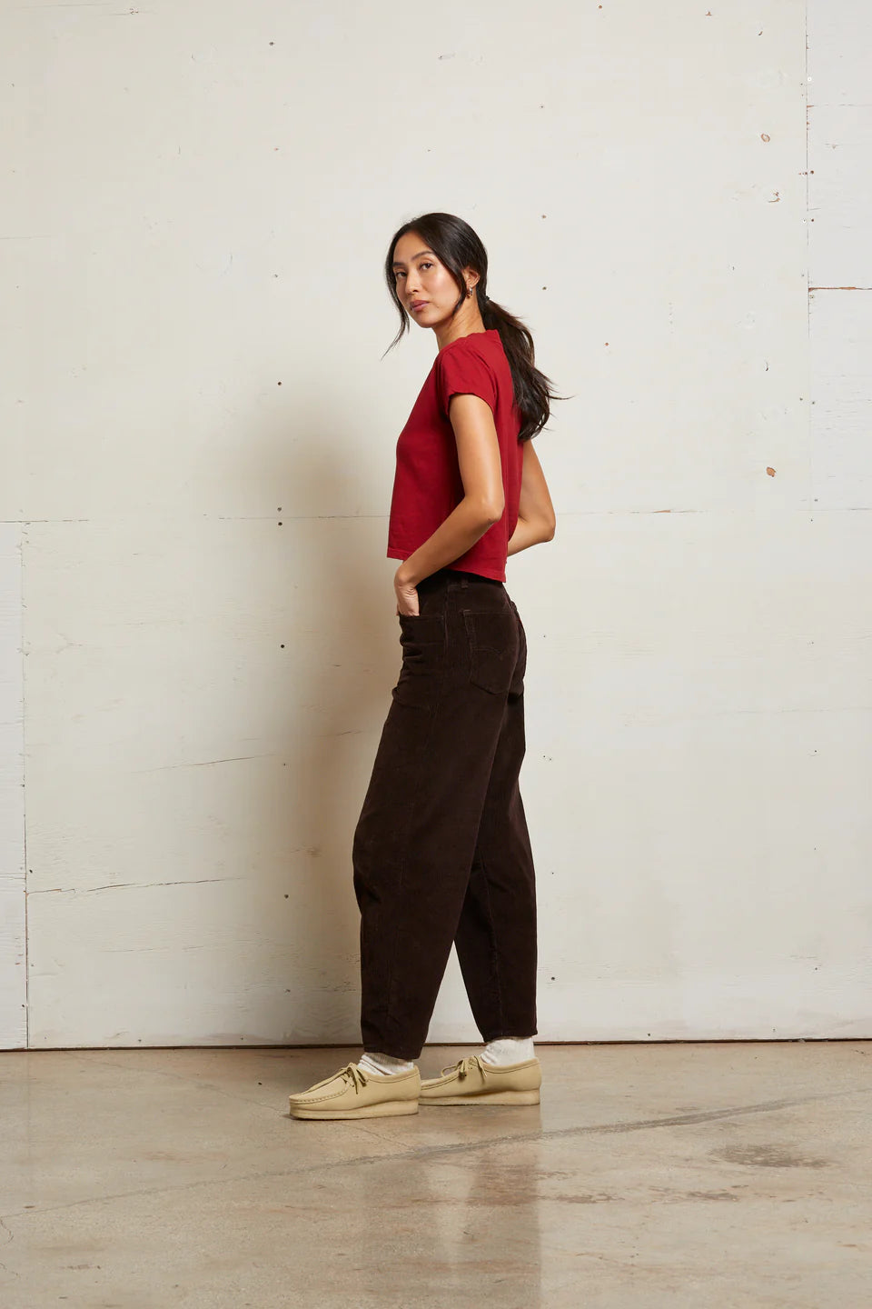 Woman wearing a red top and dark pants against a white wall side view