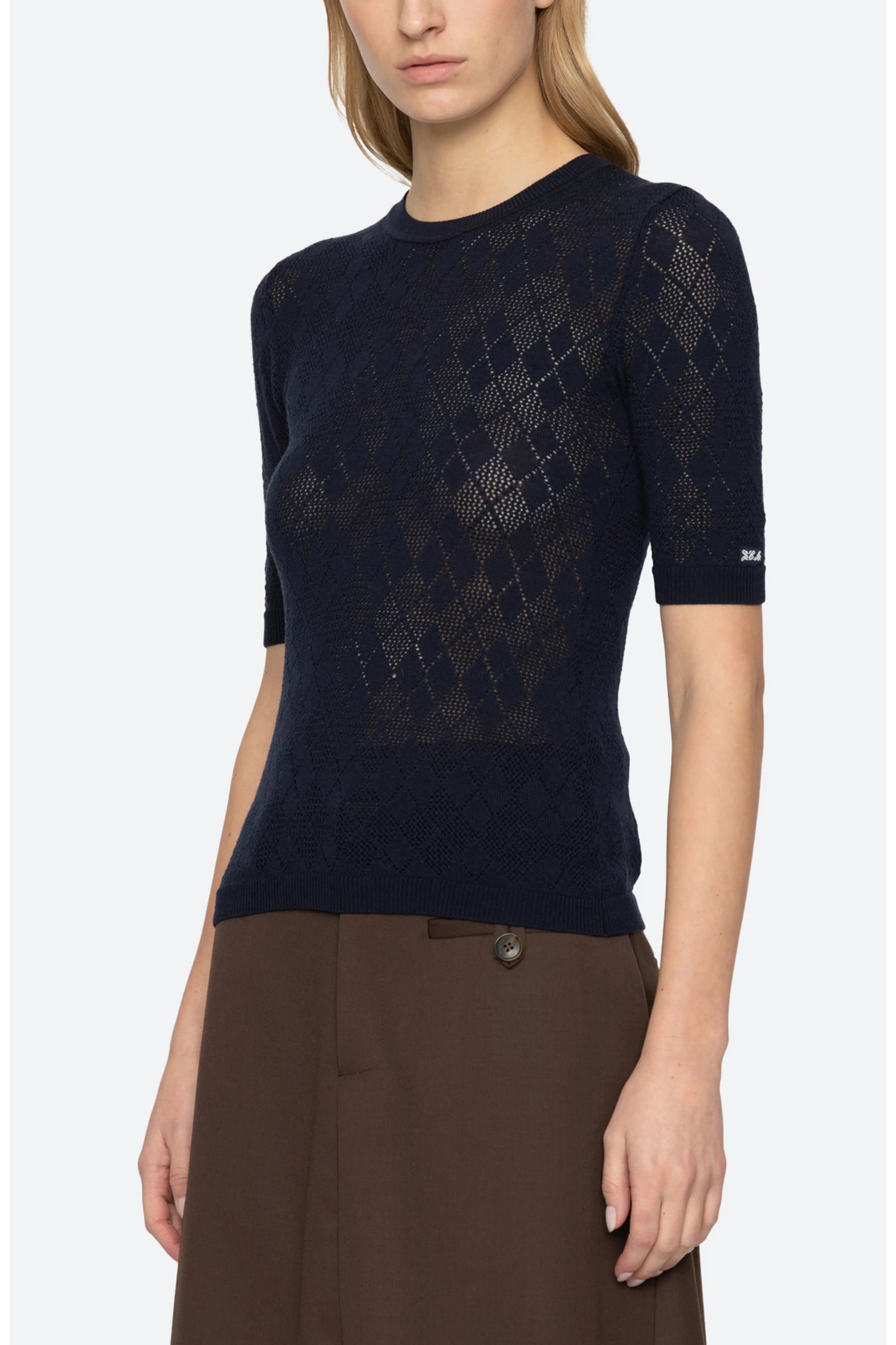 A woman wearing a dark navy blue short-sleeve knit top with a subtle argyle pattern, paired with brown trousers, against a light gray studio background.