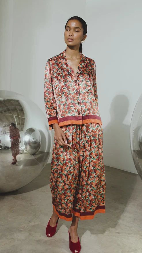 A model stands in a minimalist studio setting wearing a matching two-piece pajama set with a dense floral print in red, orange, and green tones, accented with solid orange and red stripes at the cuffs and hem.