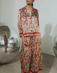 A model stands in a minimalist studio setting wearing a matching two-piece pajama set with a dense floral print in red, orange, and green tones, accented with solid orange and red stripes at the cuffs and hem.