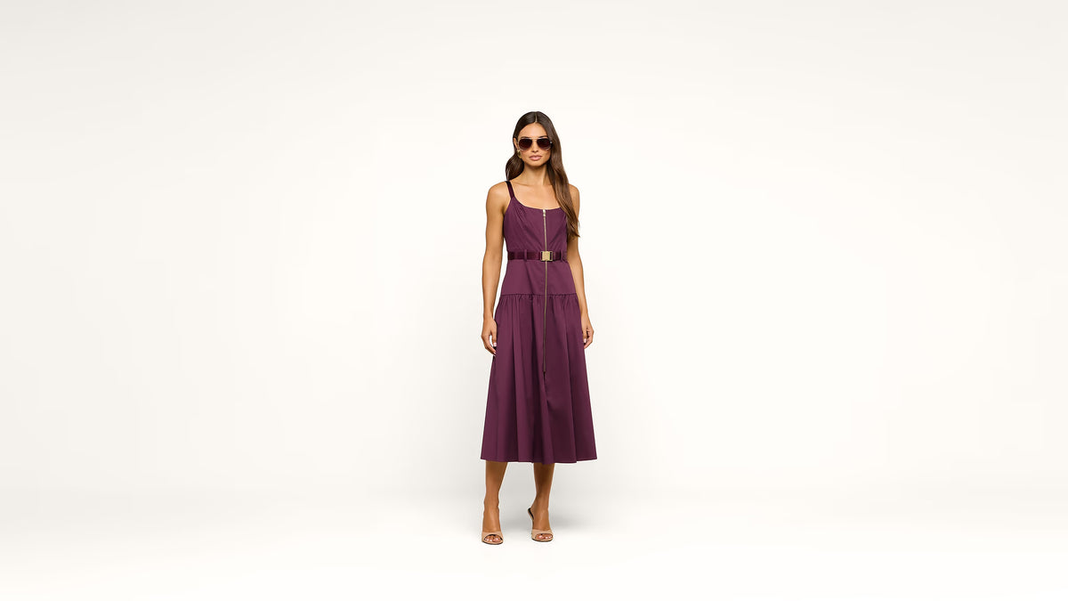 A woman modeling a sleeveless plum purple midi dress with a thin gold belt at the waist, standing against a seamless white studio background.
