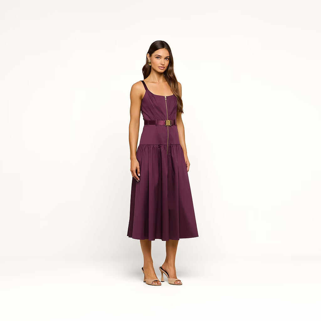 A woman modeling a sleeveless midi dress in deep plum purple with a matching waist belt and a prominent gold front zipper. She is wearing strappy high-heeled sandals against a clean, off-white studio background.
