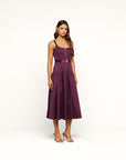 A woman modeling a sleeveless midi dress in deep plum purple with a matching waist belt and a prominent gold front zipper. She is wearing strappy high-heeled sandals against a clean, off-white studio background.