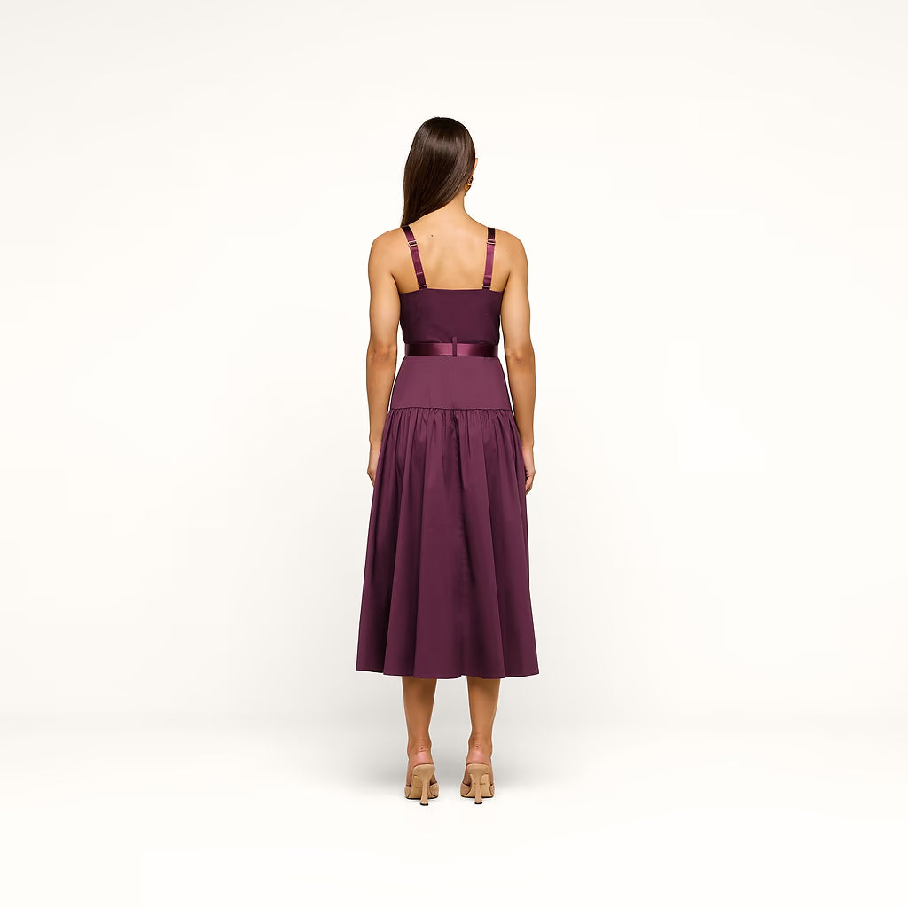 A woman viewed from the back wearing a sleeveless midi dress in deep plum purple with a matching waistband ribbon, standing against a plain white studio background.
