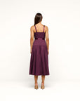 A woman viewed from the back wearing a sleeveless midi dress in deep plum purple with a matching waistband ribbon, standing against a plain white studio background.