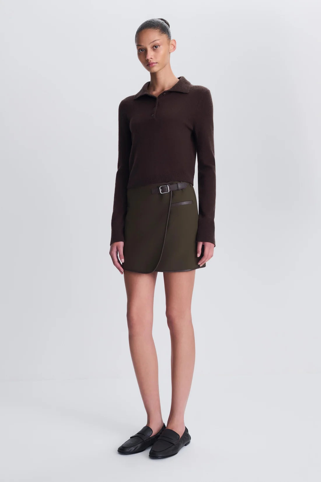 A model stands against a light gray studio background wearing a dark brown long-sleeve polo shirt, an olive green wrap-style mini skirt with a buckle detai, and black slip-on loafers.