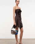 Woman wearing a dark brown strapless dress holding a colorful handbag on a plain background