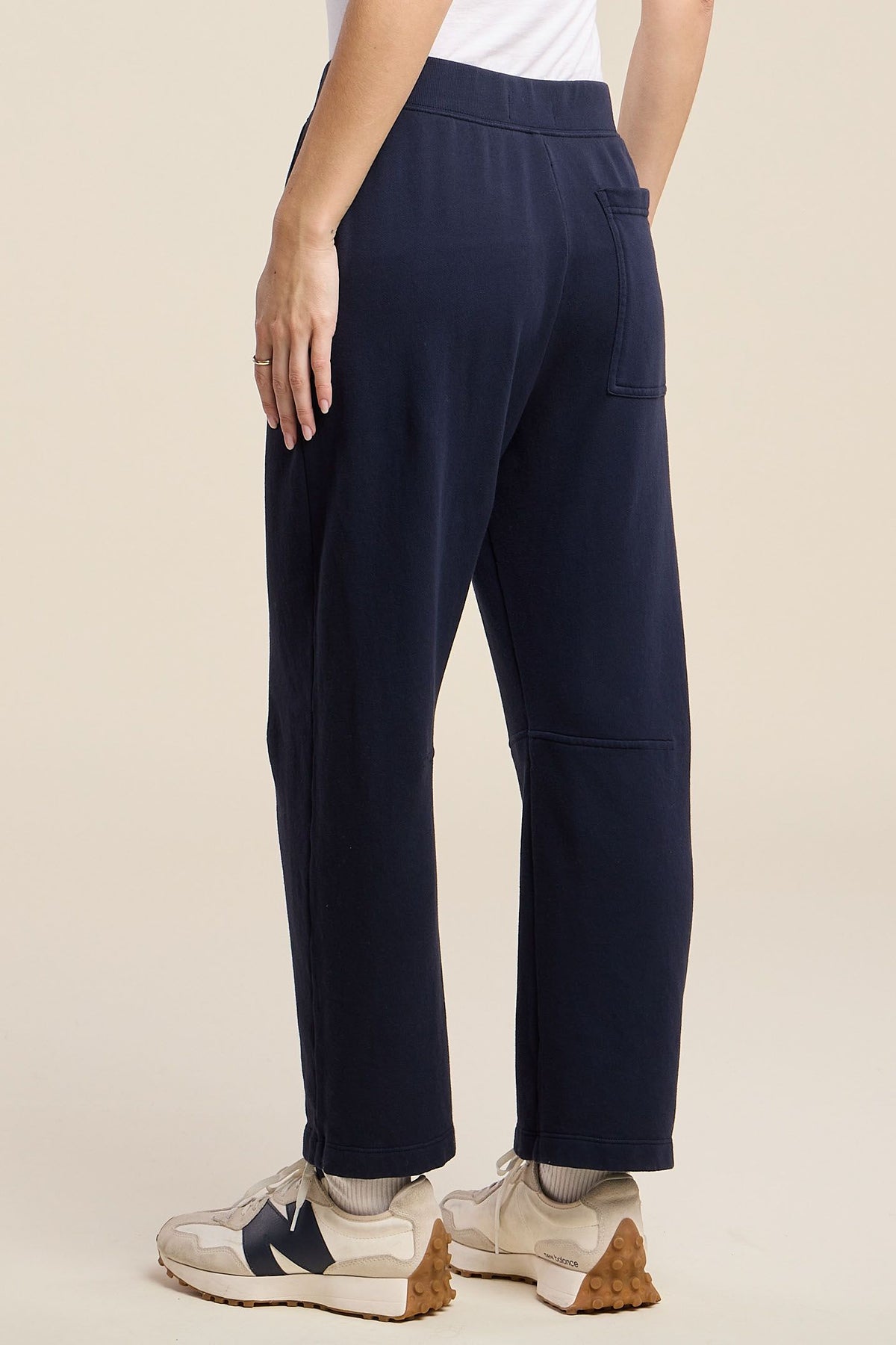 A person shown from the back wearing navy blue cropped pants with a white top and beige sneakers, photographed against a plain warm beige studio background.