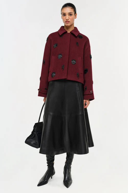 Woman wearing a burgundy coat with black embellishments and a black leather skirt on a white background