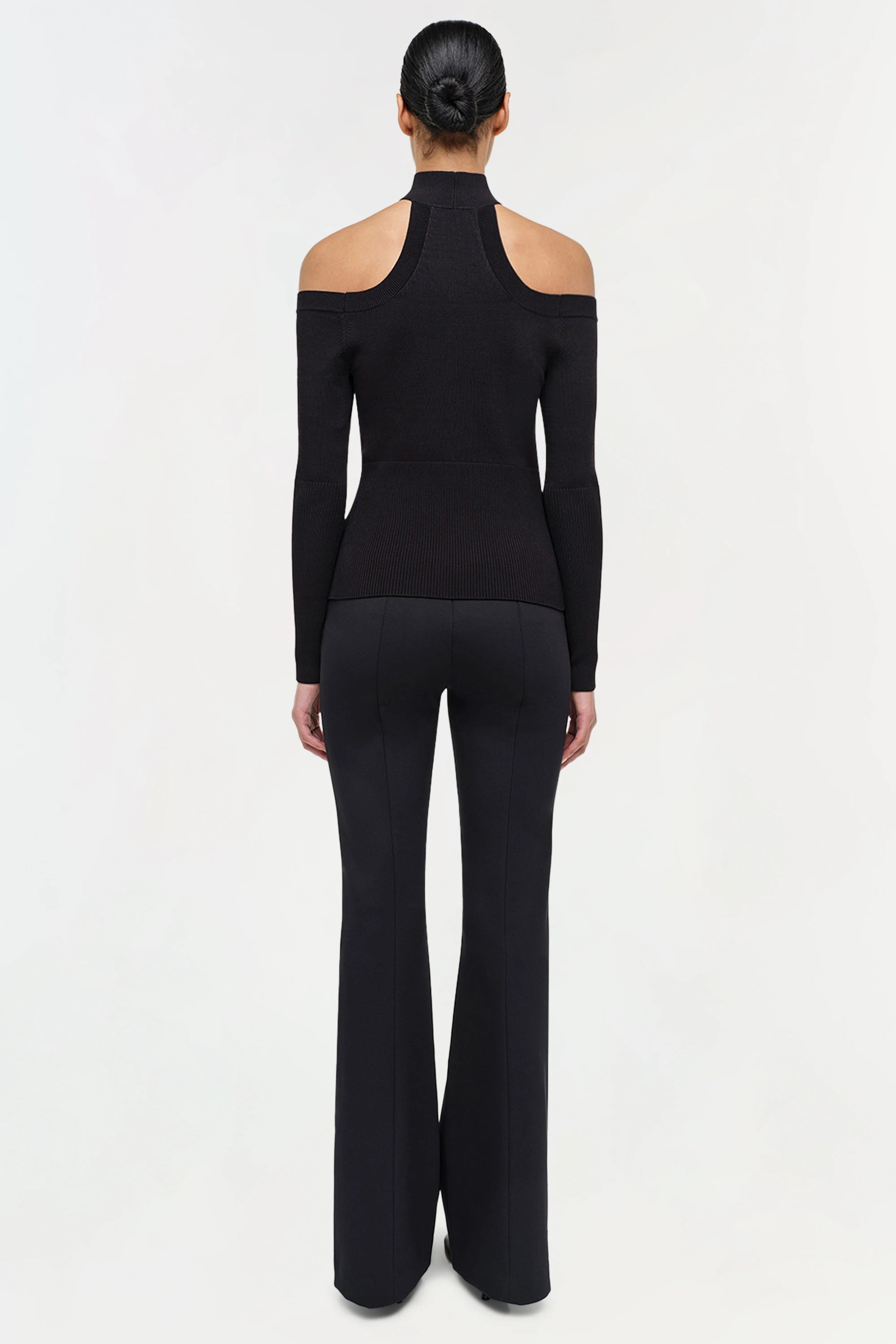 Person wearing a black outfit with a high neckline and flared pants on a white background back