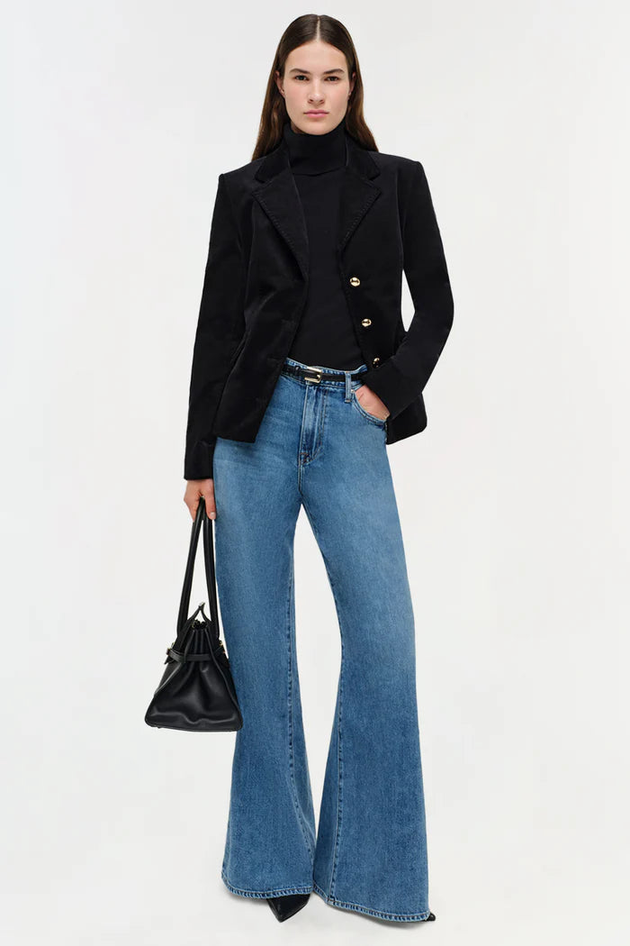 Woman wearing a black blazer, blue jeans, and holding a black handbag on a white background