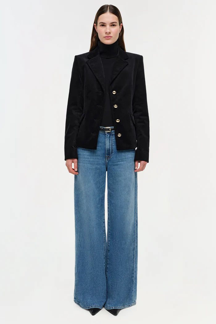 Person wearing a black blazer with gold buttons and blue jeans on a white background