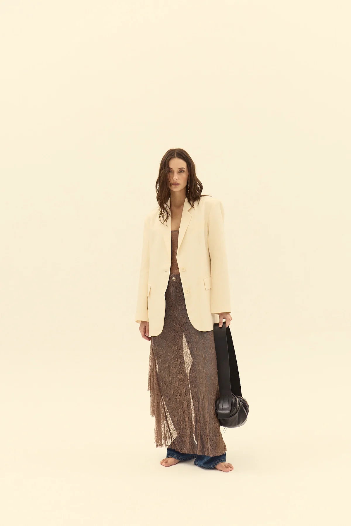 Woman wearing a beige blazer and brown dress holding a black bag on a beige background